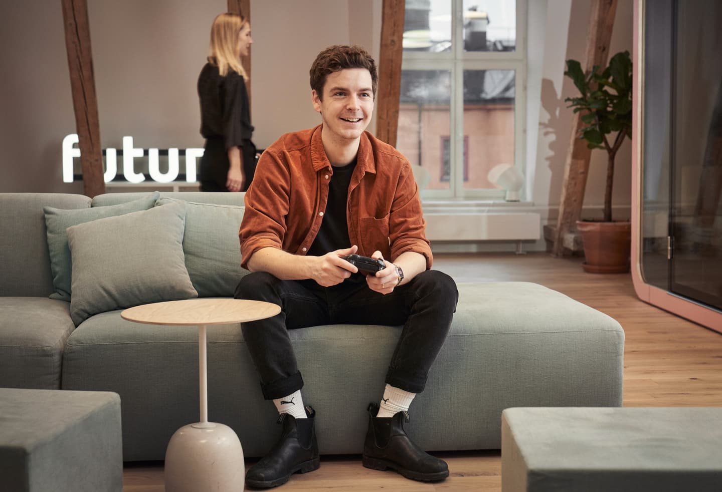 Person in orange corduroy shirt playing video games on a blue sofa in modern office with "futur" sign visible.