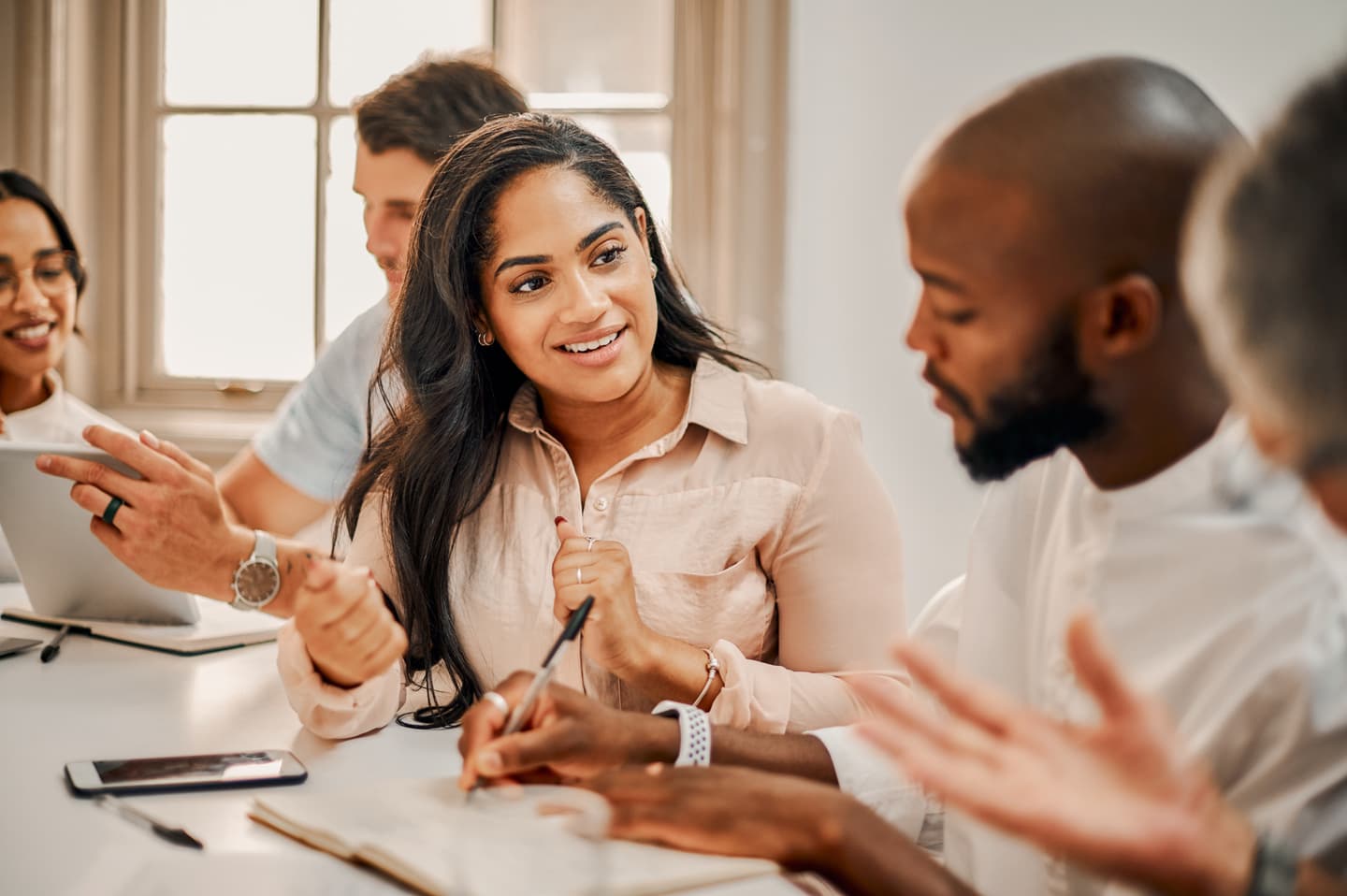 Diverse group of professionals in a bright meeting room engaged in animated discussion, with a woman in beige shirt smiling.